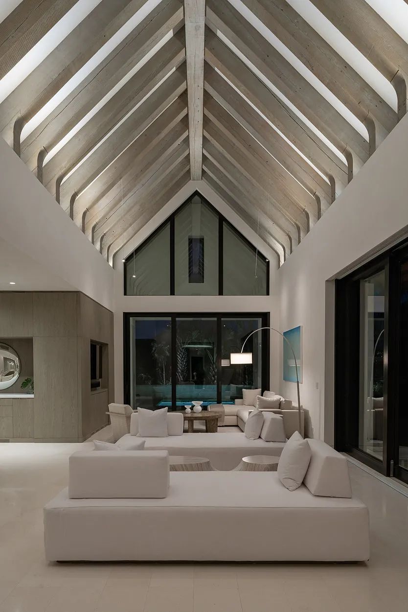 Living room with white sectional sofa, round coffee table, large windows, and exposed wooden beams.