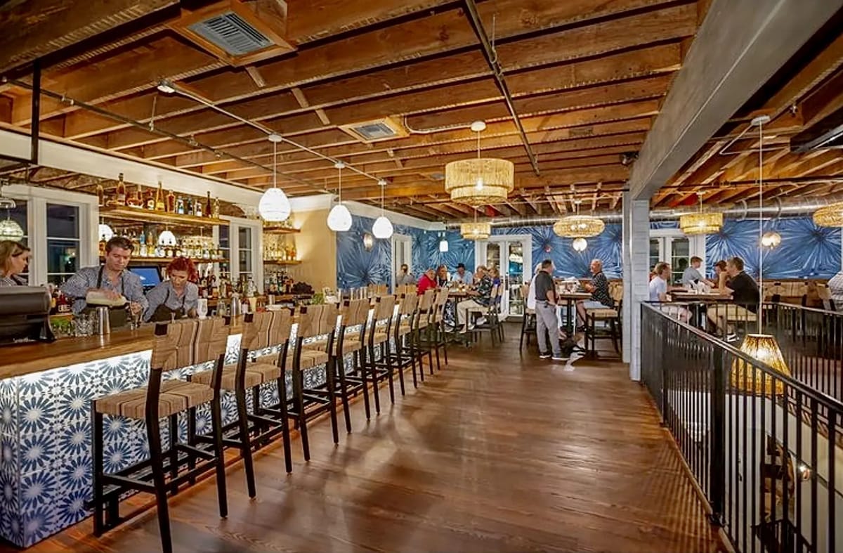 Dining room with bar, tables, chairs, wooden ceiling, and decorative lights, featuring a blue patterned wall.