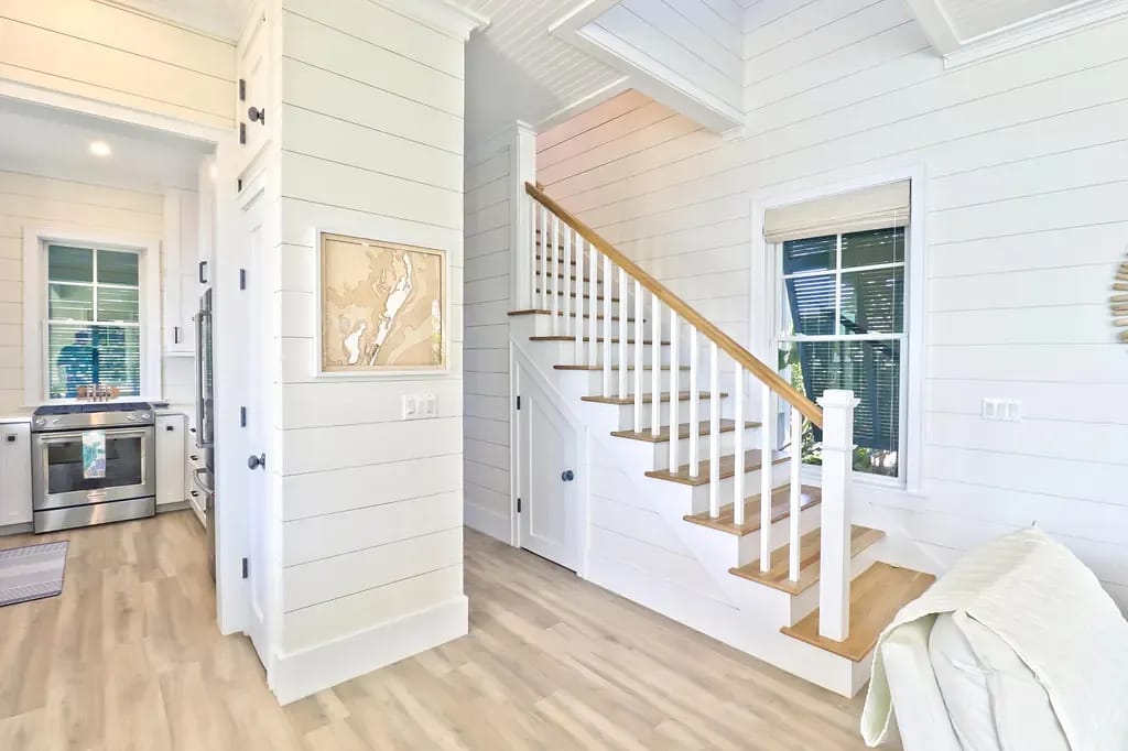 Entryway with shiplap walls, wooden staircase, and framed map leading to kitchen area.