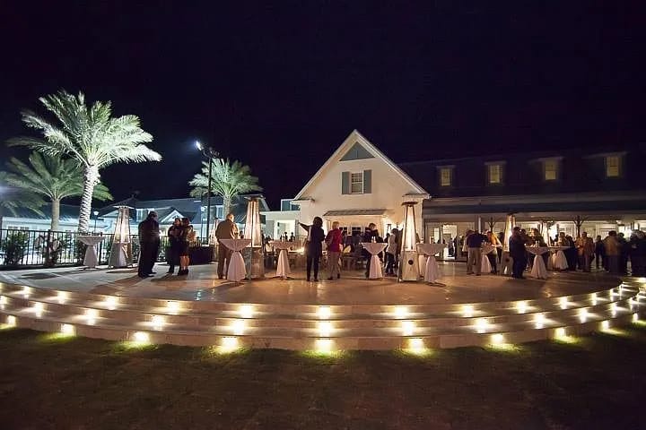 Exterior rear area with round dining tables, palm trees, and a large white house at night with soft lighting.