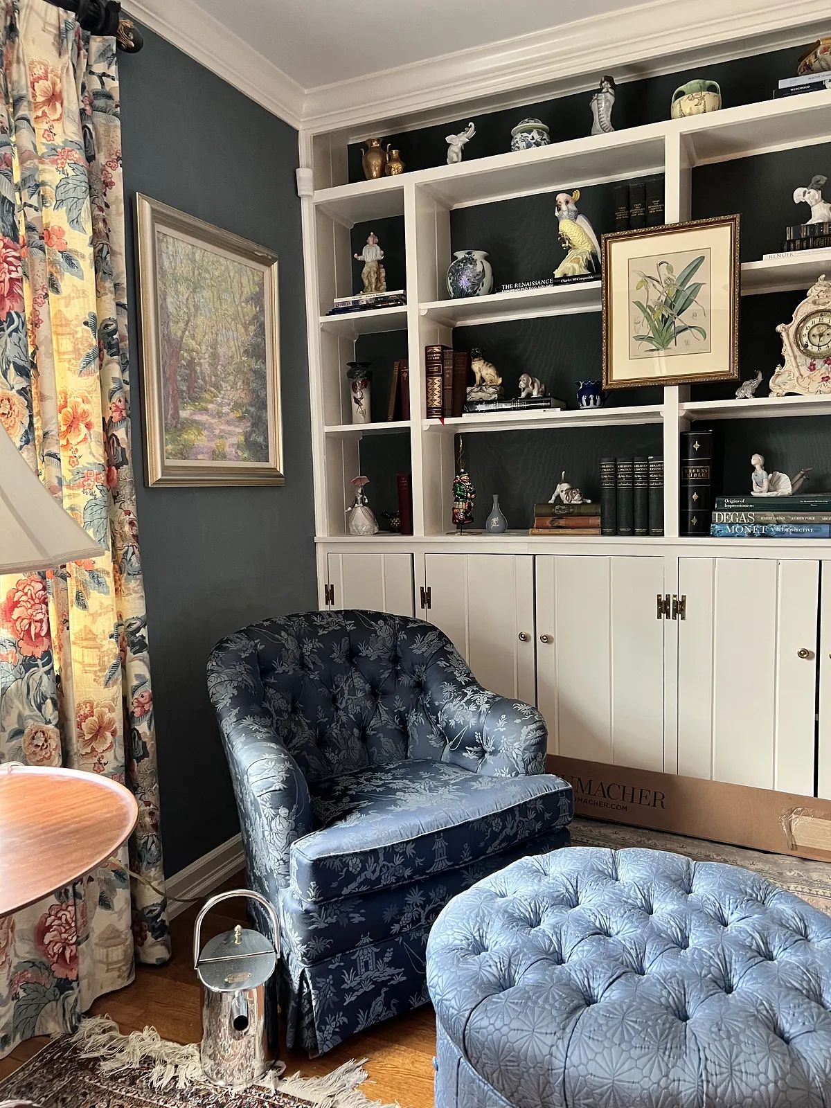 Living room with blue armchair, round ottoman, side table, built-in bookshelf, and floral curtains on dark blue walls