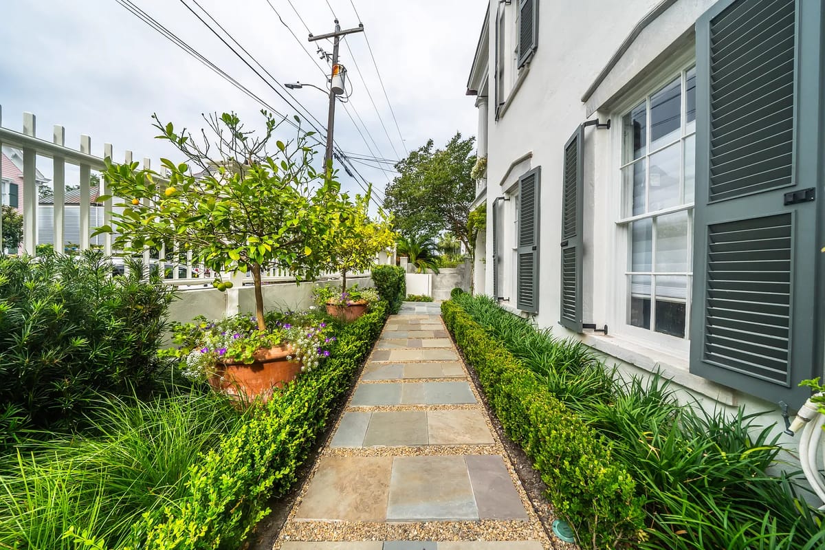 Garden pathway lined with shrubbery and potted plants, featuring stone slabs and a white fence.