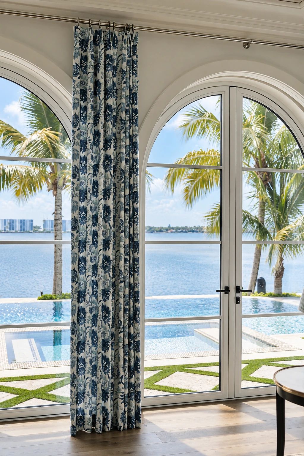 View with large arched windows, patterned curtains, palm trees, and a pool outside.
