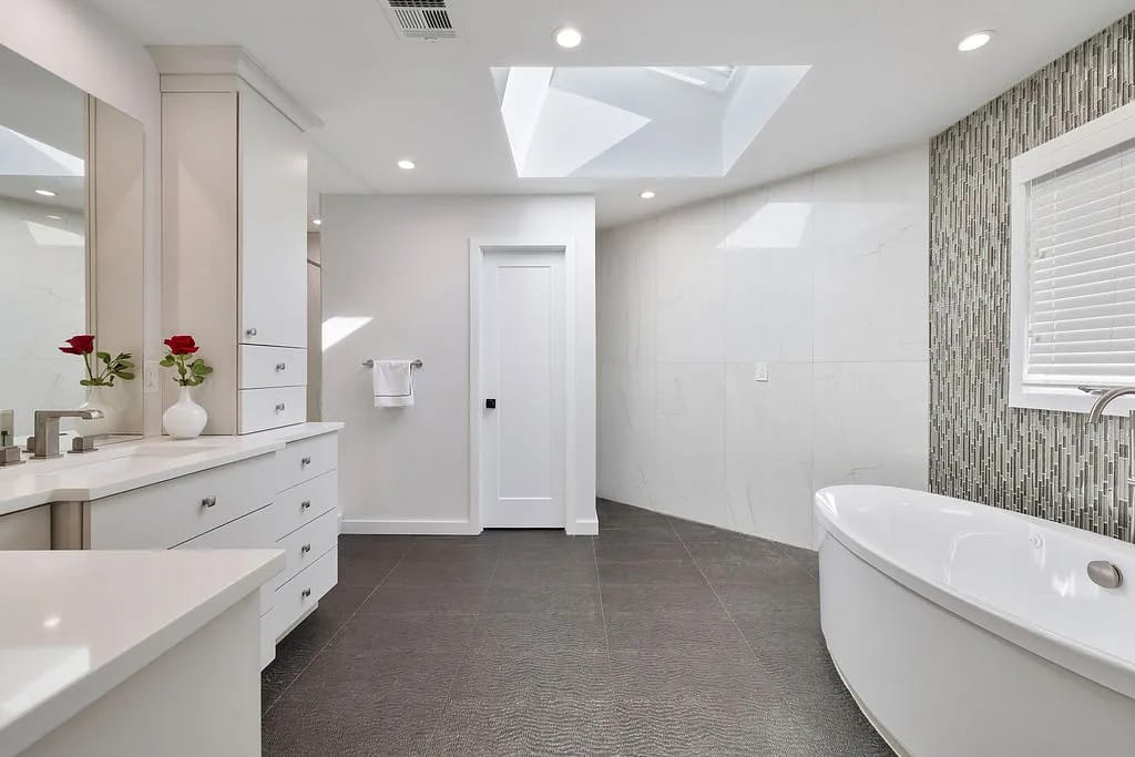 Bathroom with freestanding white bathtub, large vanity, dual sinks, grey tile floor, and skylight.