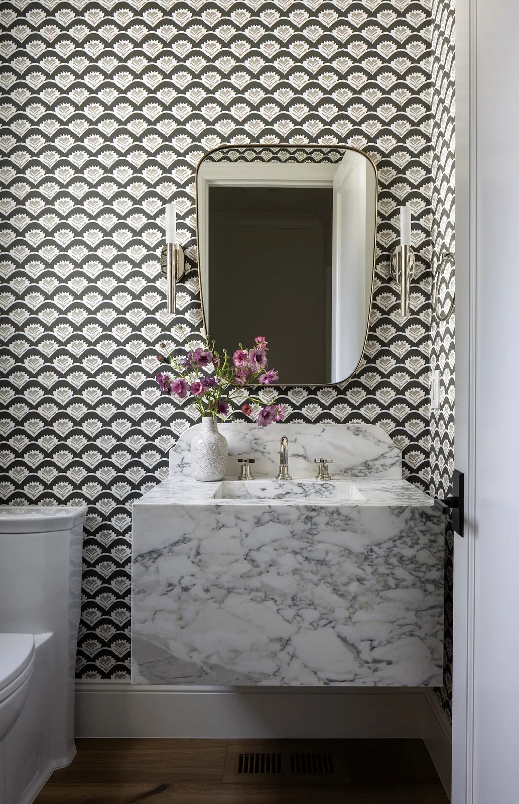 Bathroom with marble sink, patterned wallpaper, decorative mirror, toilet, and small vase with flowers.