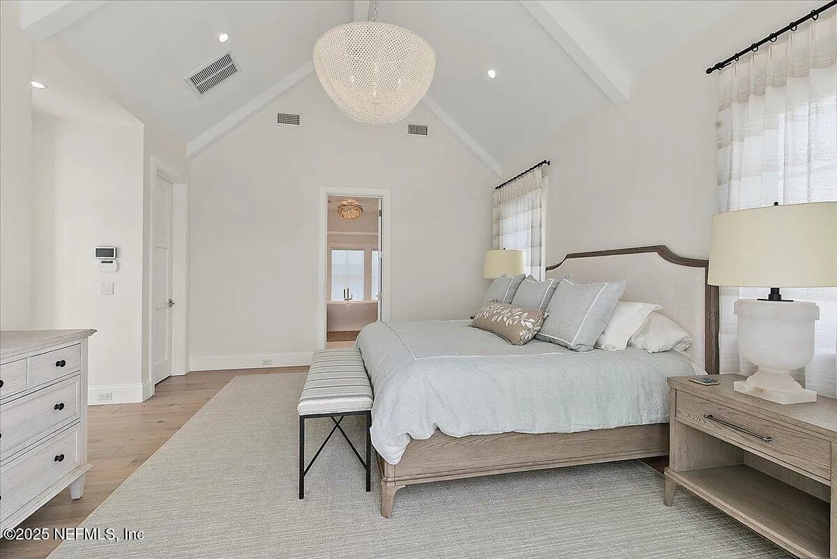 Bedroom with gray bed, wooden nightstand, chest of drawers, bench, and large windows with sheer curtains