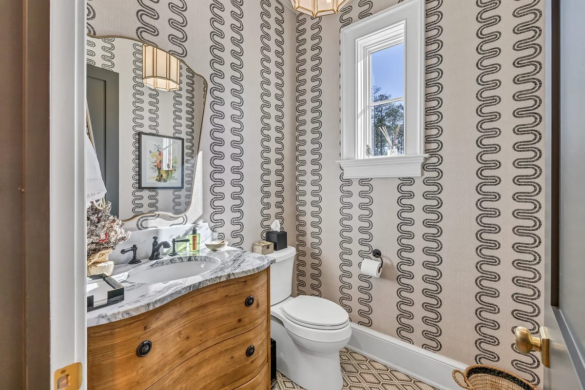 Powder room with wooden vanity, sink, toilet, round mirror, patterned wallpaper, and a small window.
