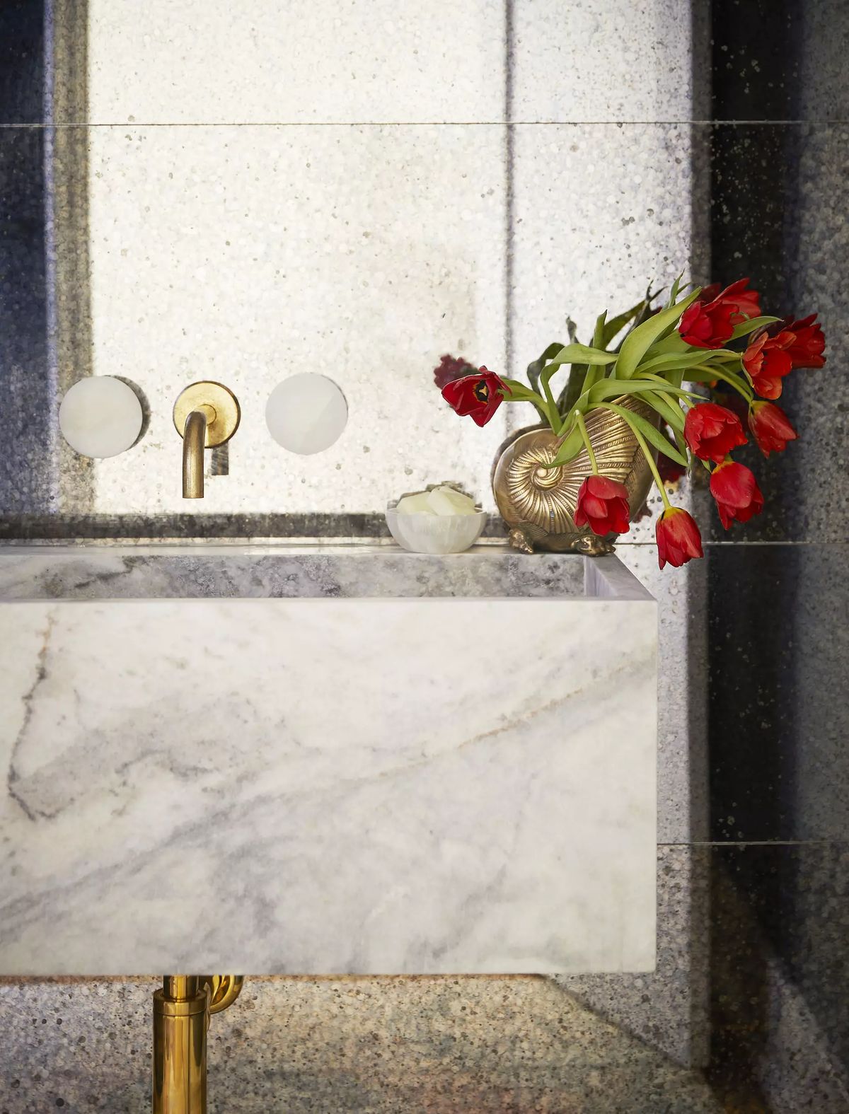 Bathroom with marble sink, gold faucet, wall-mounted fixtures, and vase of red tulips.