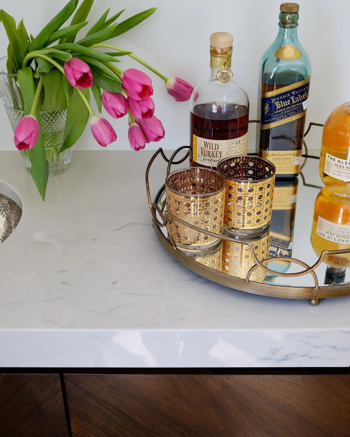 Countertop with marble, pink tulips, decorative glasses, and three liquor bottles on a tray.