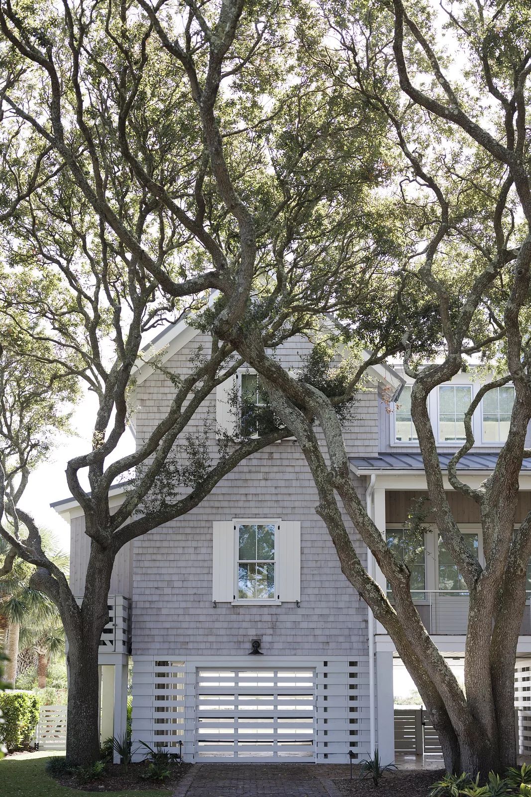 Exterior front of a house with shingle siding, trees, driveway, white shutters, and garage door.