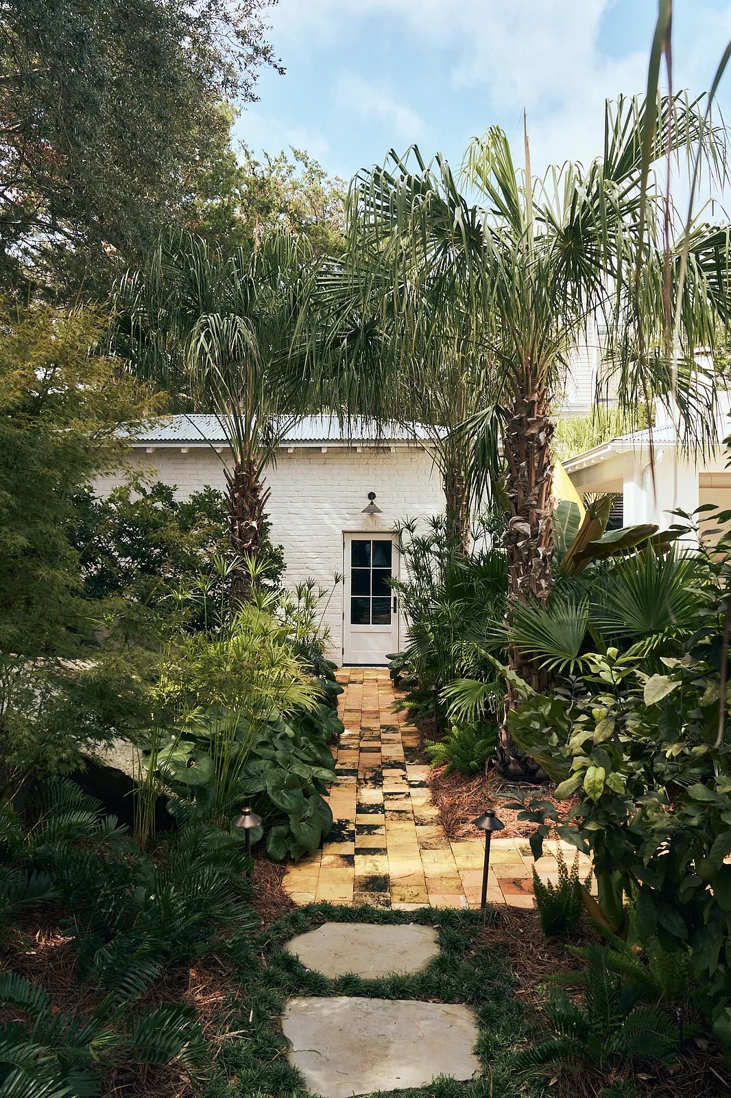 Garden with stone pathway, palm trees, shrubs, and a white brick building with a black door.
