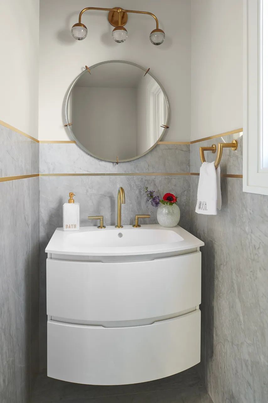 Bathroom with white vanity, gold faucets, round mirror, grey marble backsplash, and flowers in a vase.