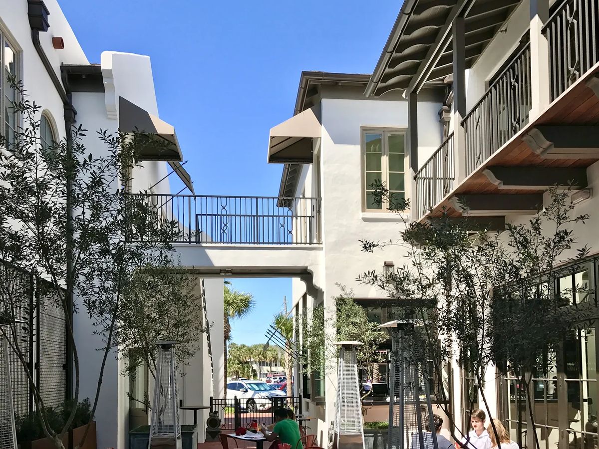 Outdoor area with two buildings, walkway, olive trees, outdoor heaters, and metal furniture under a blue sky.