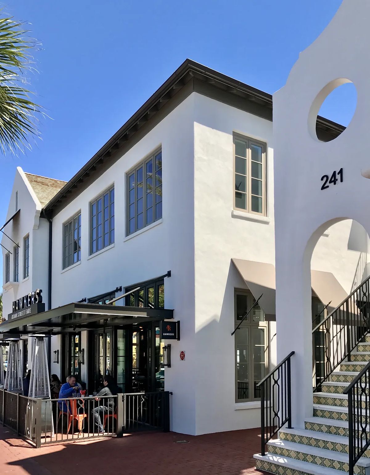 Exterior front of a white stucco building with large windows, staircase, and outdoor seating area.