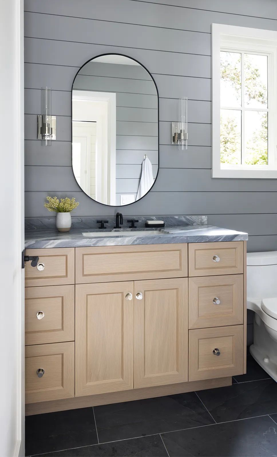 Bathroom with light wood vanity, marble countertop, circular mirror, wall sconces, and gray shiplap paneling.