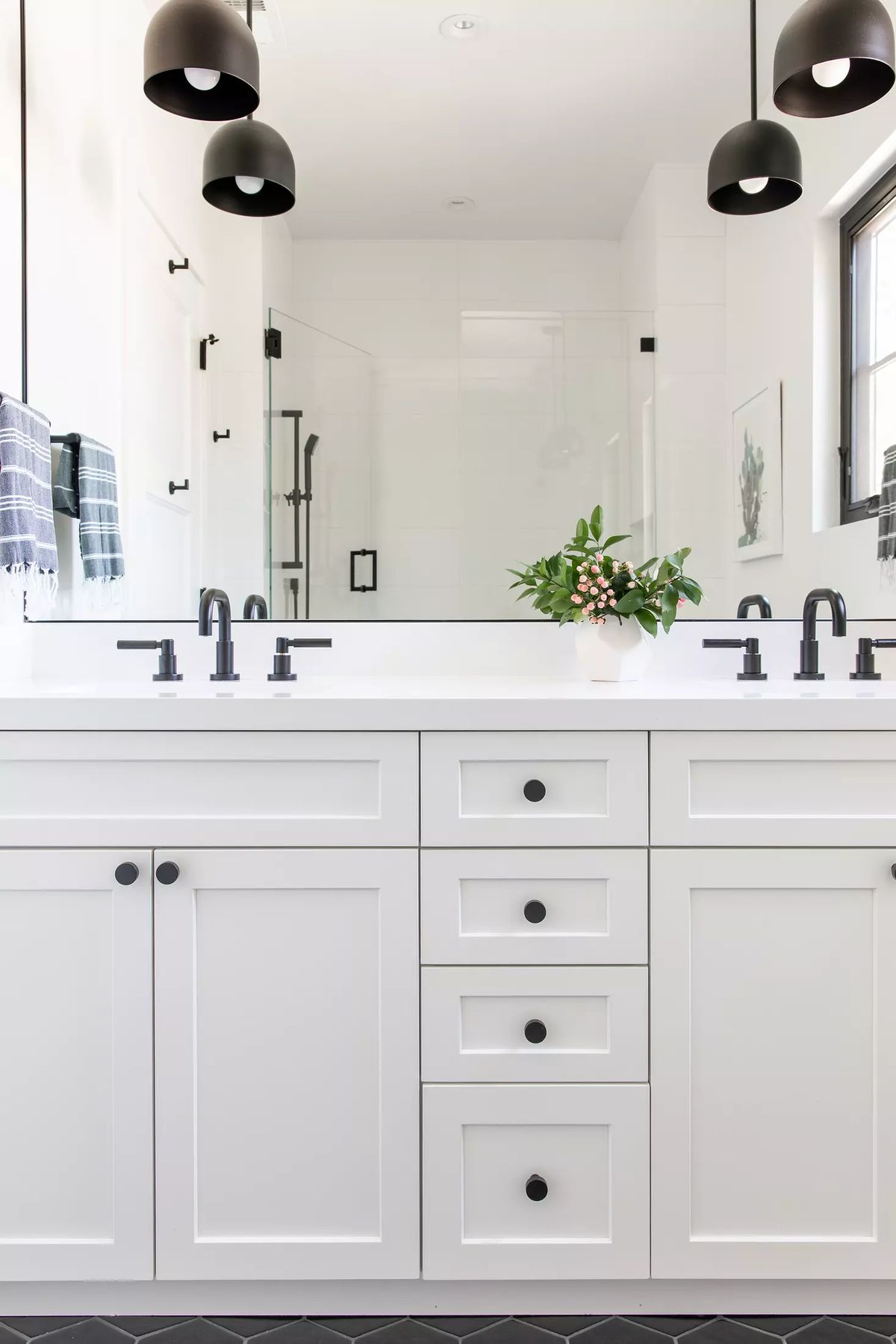 Bathroom with white vanity, black pendant lights, gray towels, and glass-enclosed shower on herringbone tile floor.