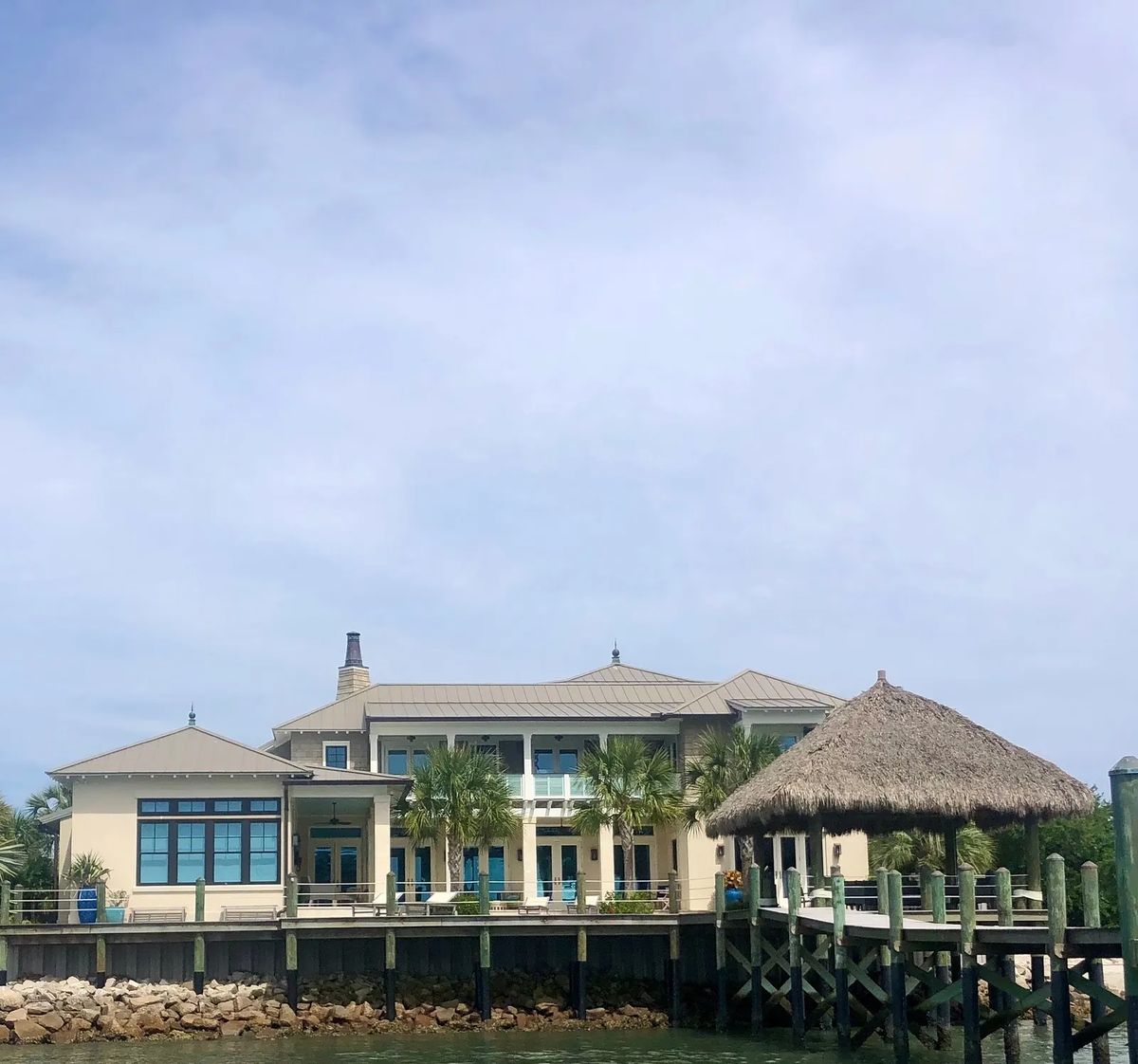 Exterior view of a house with patio, dock, palm trees, and thatched-roof structure.