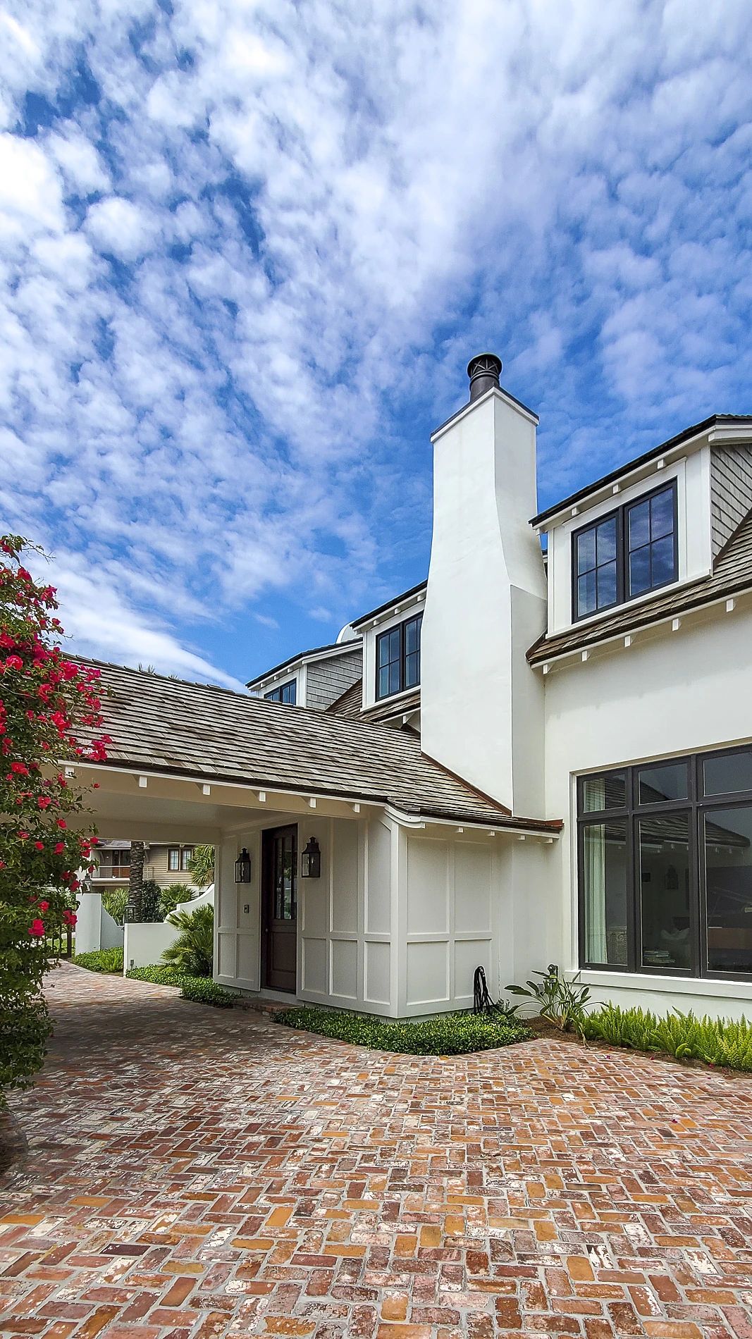 Exterior front view with brick driveway, white stucco walls, chimney, large windows, and flowering bush
