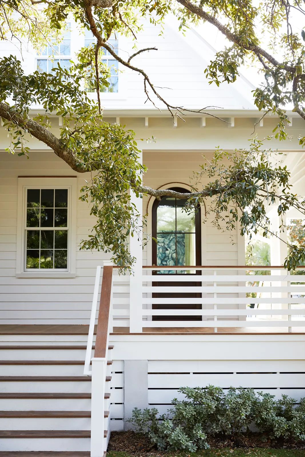 Exterior front of a house with white facade, staircase, arched door, and tree branch overhead