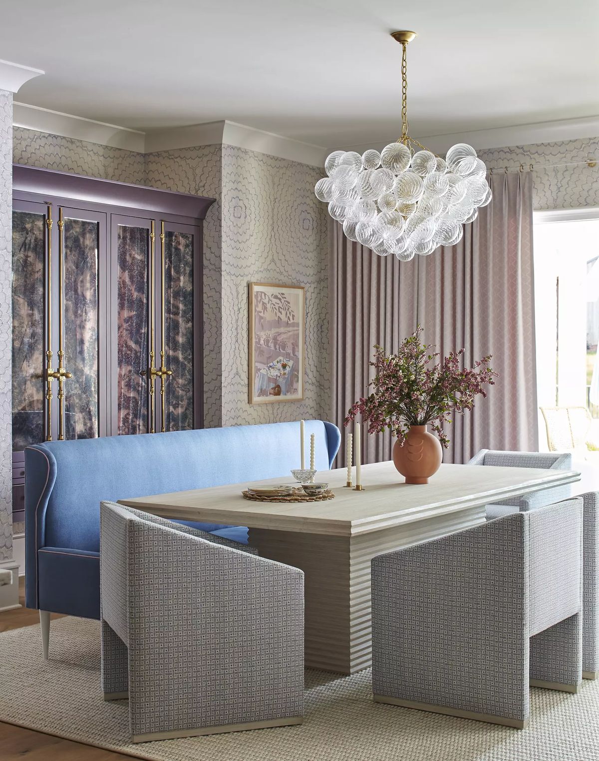 Dining room with wooden table, blue banquette, gray chairs, floral arrangement, and patterned wallpaper.