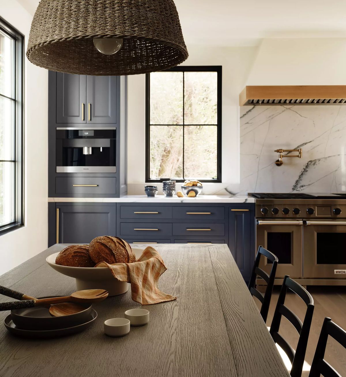 Kitchen with dark blue cabinetry, marble backsplash, large stove, wooden table, bowl of bread, and natural light from windows.