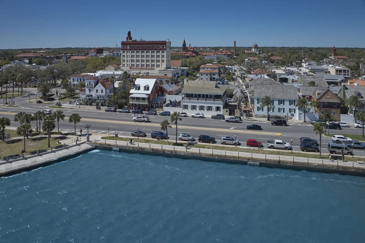 Coastal view with waterfront, highway, parked cars, palm trees, and skyline of buildings.