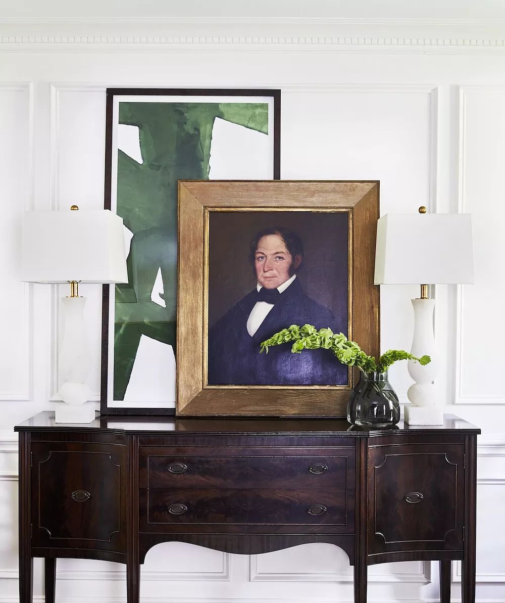 Sideboard with portrait, lamps, abstract artwork, and vase with green foliage arrangement.