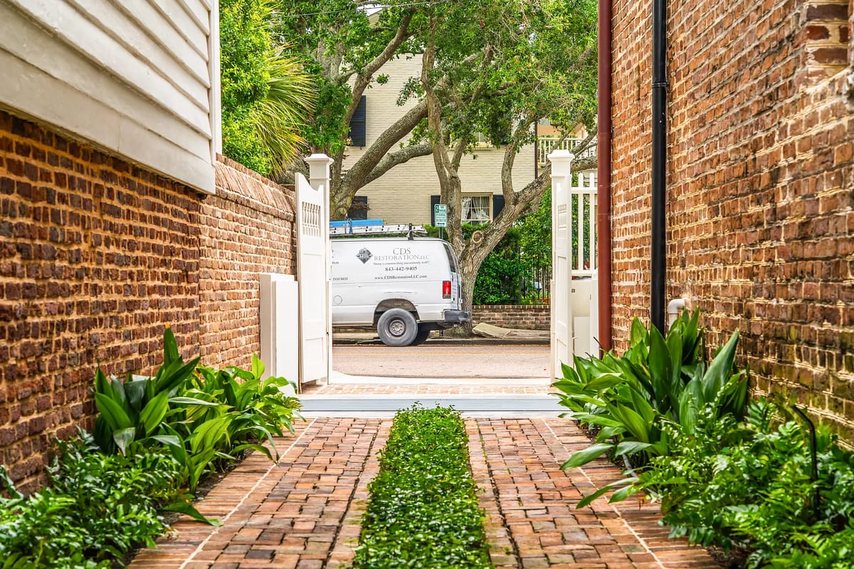 Pathway lined with greenery, leading to a white gate, surrounded by brick walls.