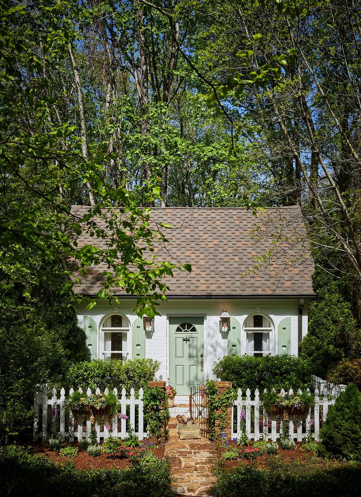 Exterior front of a cottage with green door, windows, picket fence, and stone walkway surrounded by greenery.