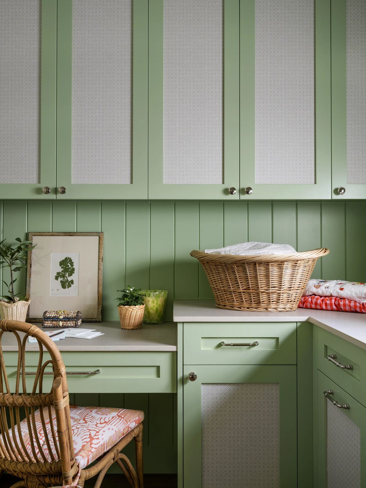 Laundry room with green cabinetry, countertop, wicker basket, framed print, chair, and potted plant.