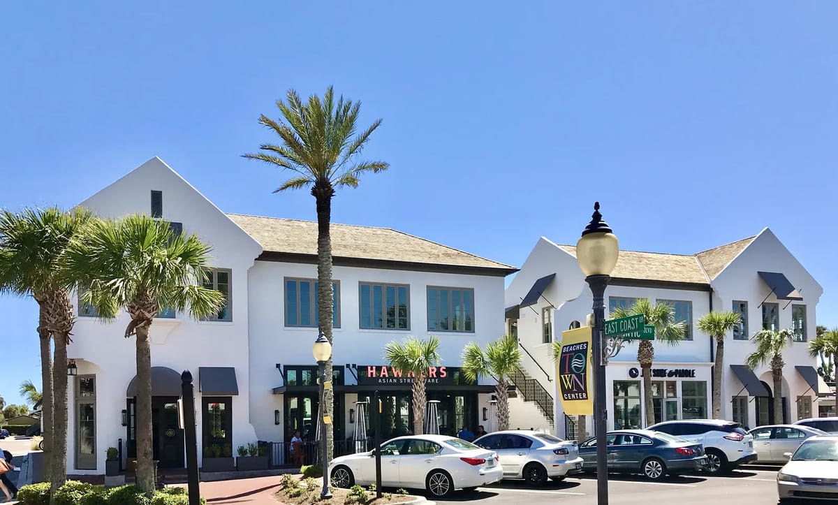 Exterior view of a two-story commercial building with white facade, wood accents, palm trees, and parked cars.