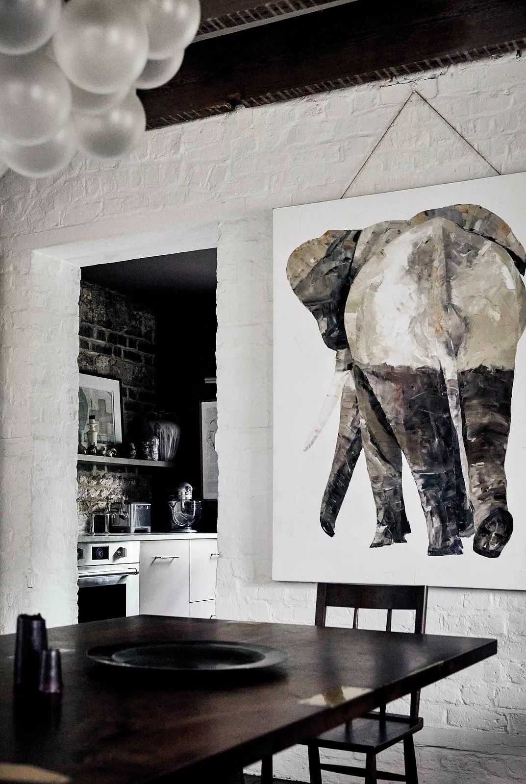 Dining room with wooden table, chairs, elephant painting, exposed brick, and kitchenette in the background.