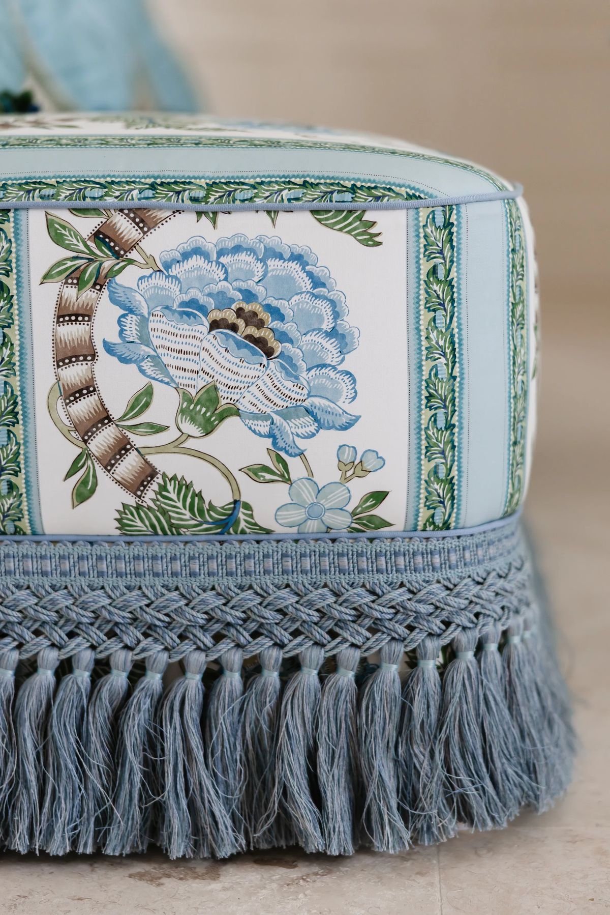 Decorative ottoman with floral pattern in blue and green, featuring light blue braided fringe.