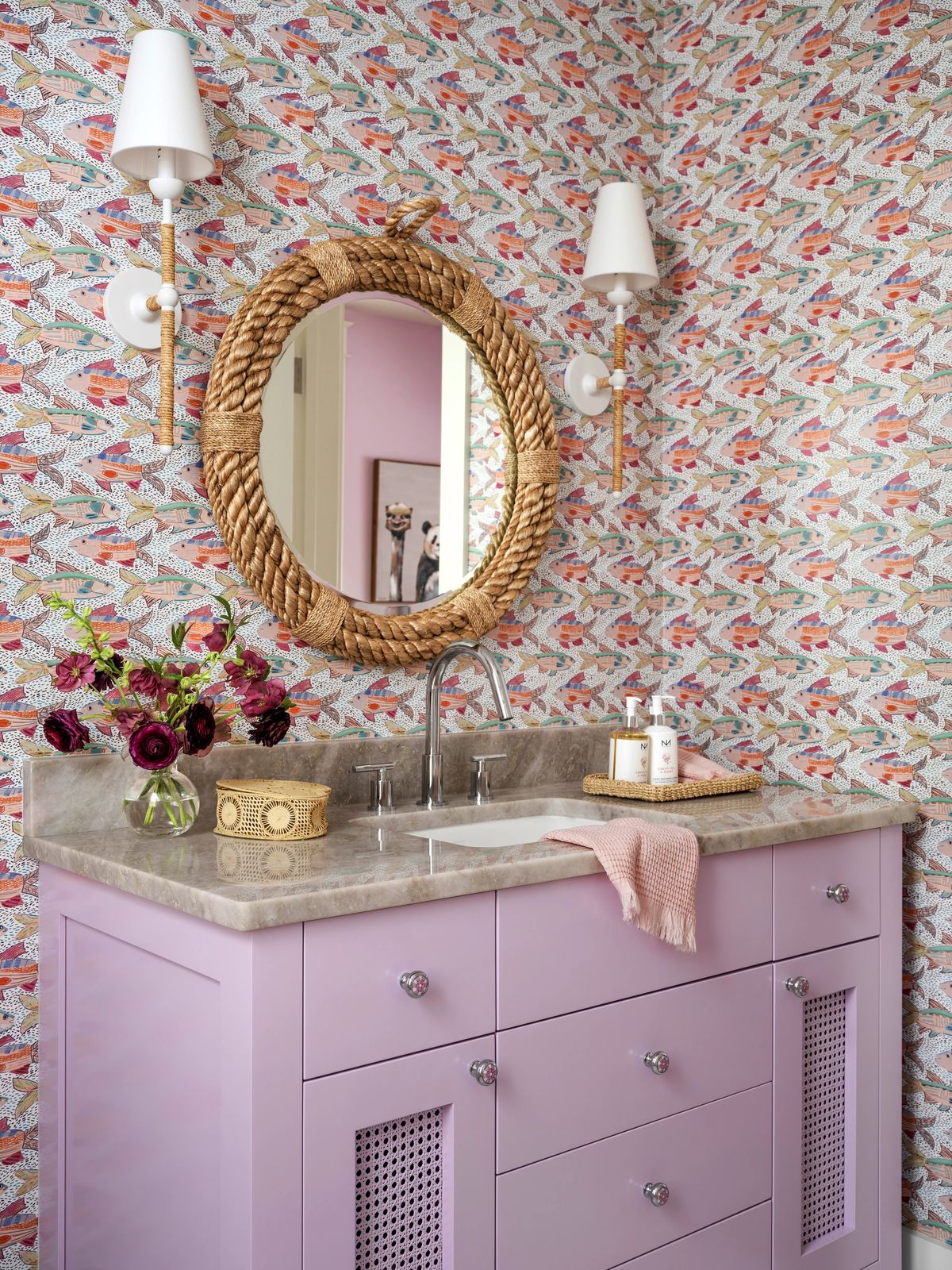 Bathroom with lavender vanity, gray countertop, patterned wallpaper, and round woven mirror.