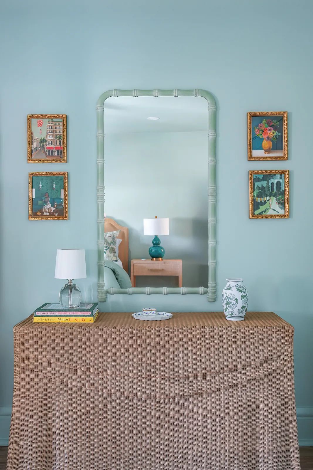 Bedroom area with console table, lamp, vase, books, mirror, and framed artworks on light blue wall.