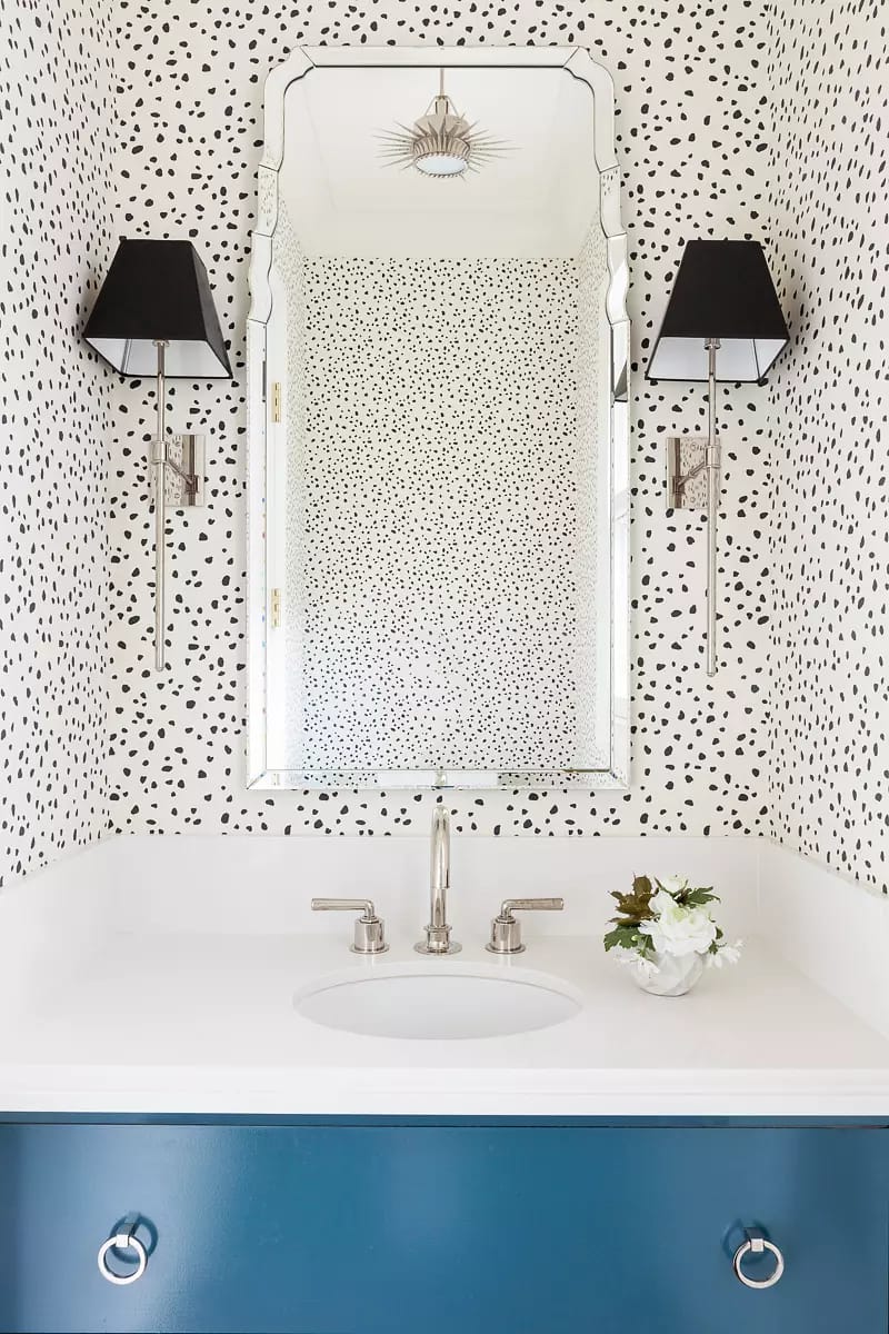 Powder room with white sink, blue cabinet, mirrored wall, and speckled black and white wallpaper.