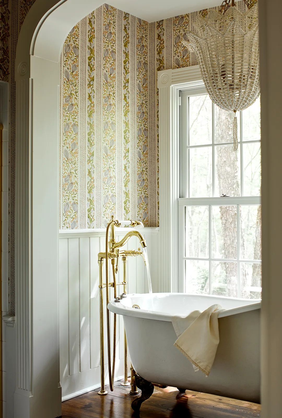 Bathroom with clawfoot tub, brass faucet, floral wallpaper, large window, and dark wood floor.