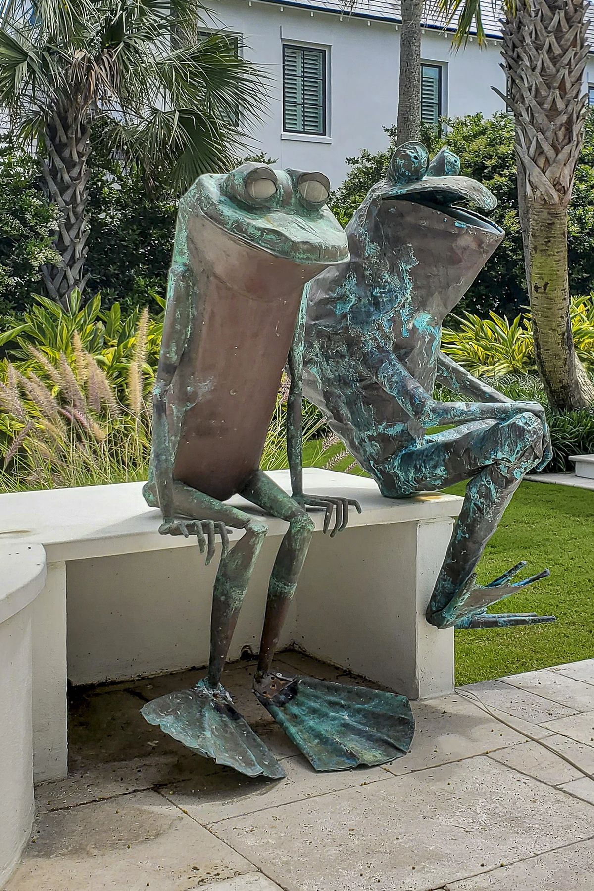 Garden area with two bronze frog sculptures on a stone base amidst greenery and palm trees.