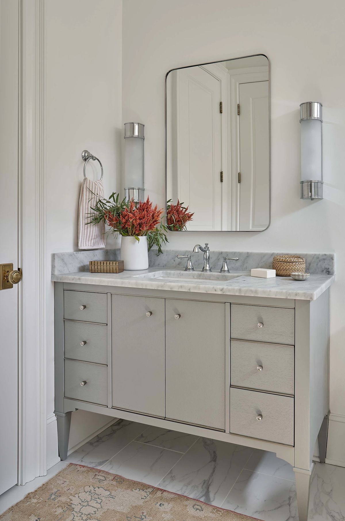 Bathroom with double vanity, marble countertop, faucet, mirror, sconces, and floral arrangement on the counter.