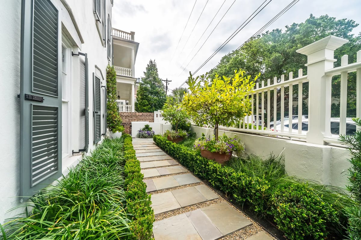 Outdoor garden with stone pathway, hedges, potted plants, and a white picket fence.