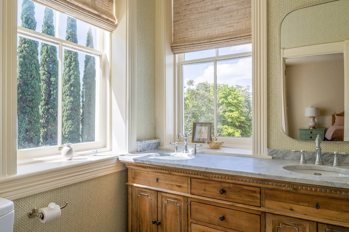Bathroom with wooden vanity, marble countertop, dual sinks, large window, and textured wallpaper.