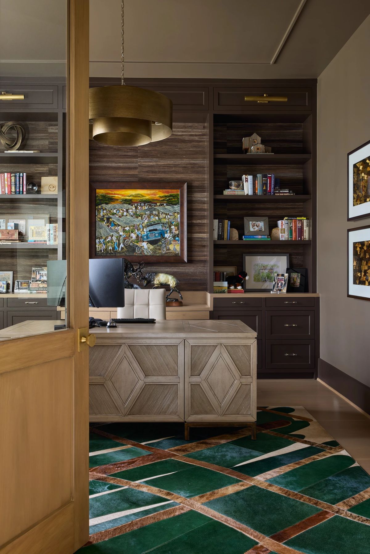 Office with wooden desk, computer, bookshelves, large artwork, and green area rug on hardwood flooring