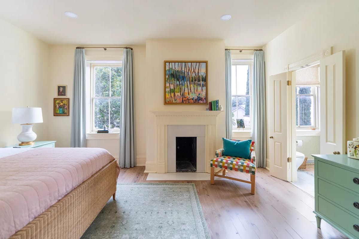 Bedroom with beige bed, pink quilt, turquoise chair, fireplace, and large windows with blue curtains.