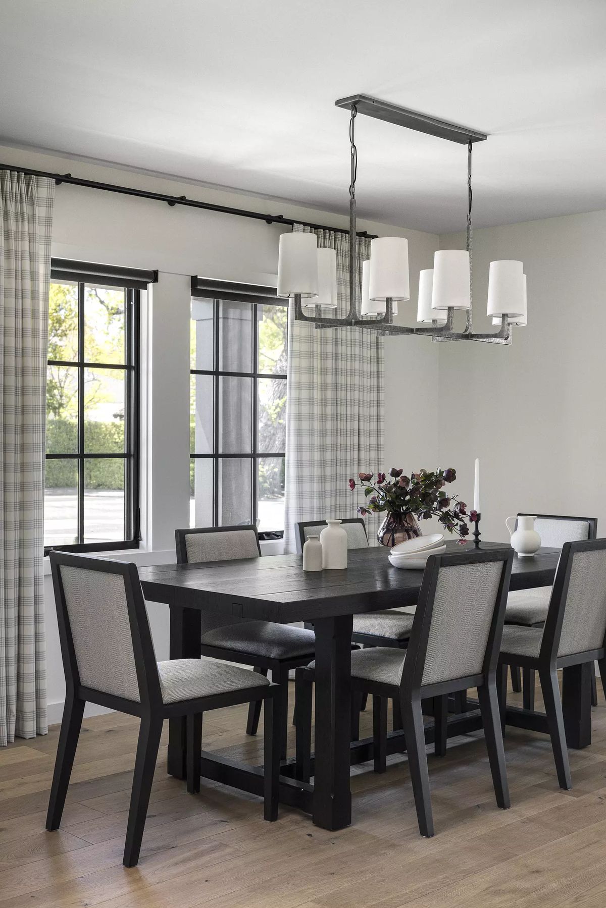 Dining room with black table, six upholstered chairs, two windows, and floral centerpiece.
