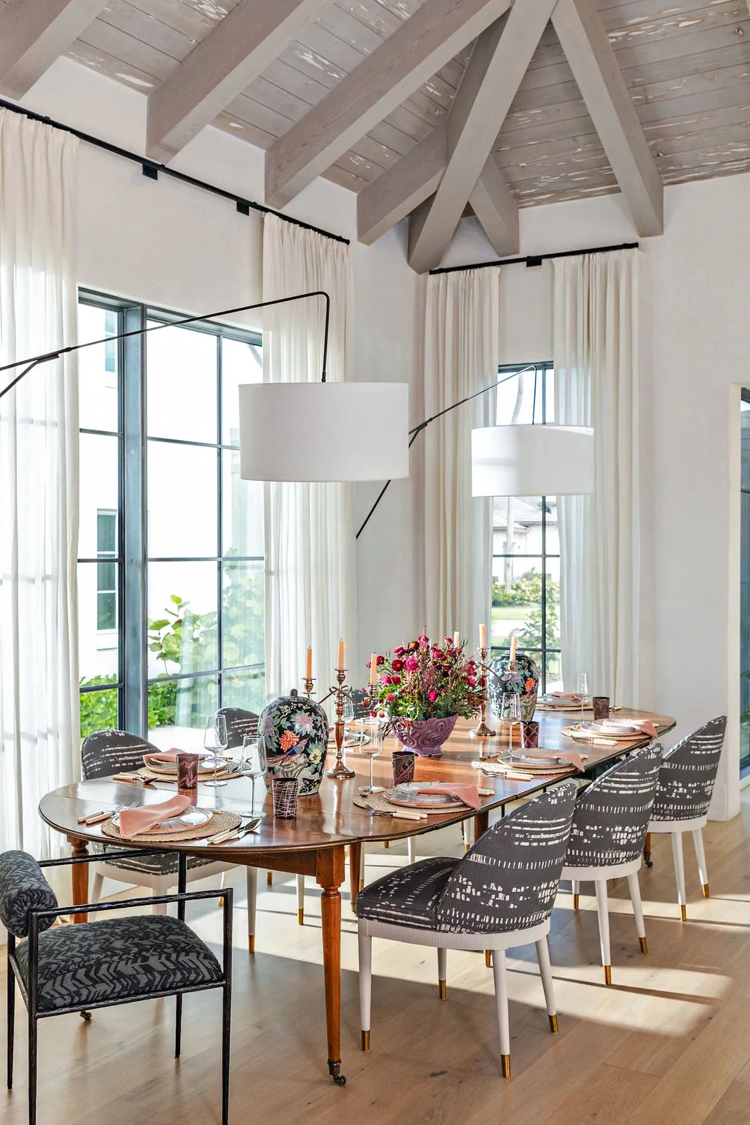 Dining room with round wooden table, patterned chairs, flowers, large windows, and pendant lights.