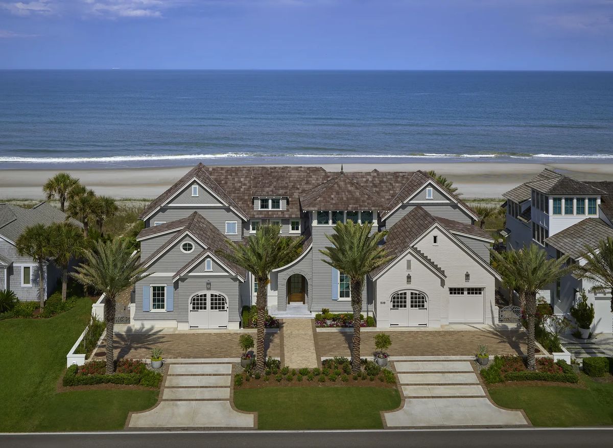 Front exterior view of a large home with gray siding, arches, large windows, palm trees, and brick paver driveway.