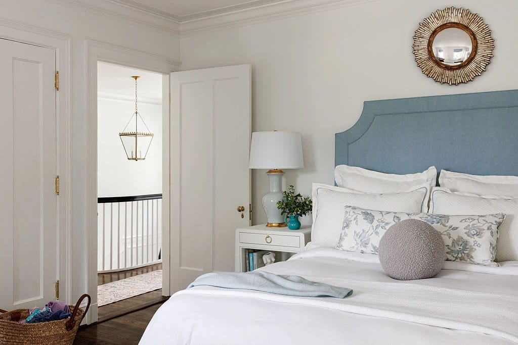 Bedroom with blue headboard, white bed linens, nightstand with lamp, and round mirror on the wall.