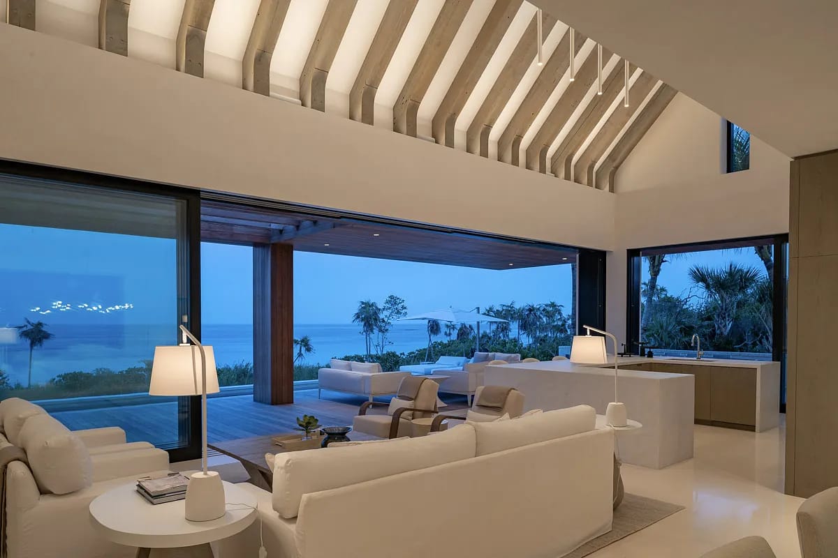 Living room with white sofas, coffee table, large windows, wooden beams, and kitchen island in the background.