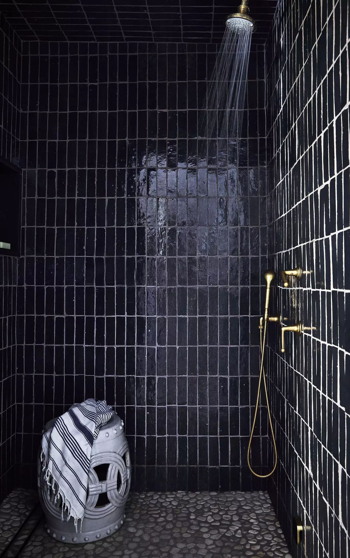 Bathroom with black tile walls, gold fixtures, pebble floor, and a stool with a striped towel.