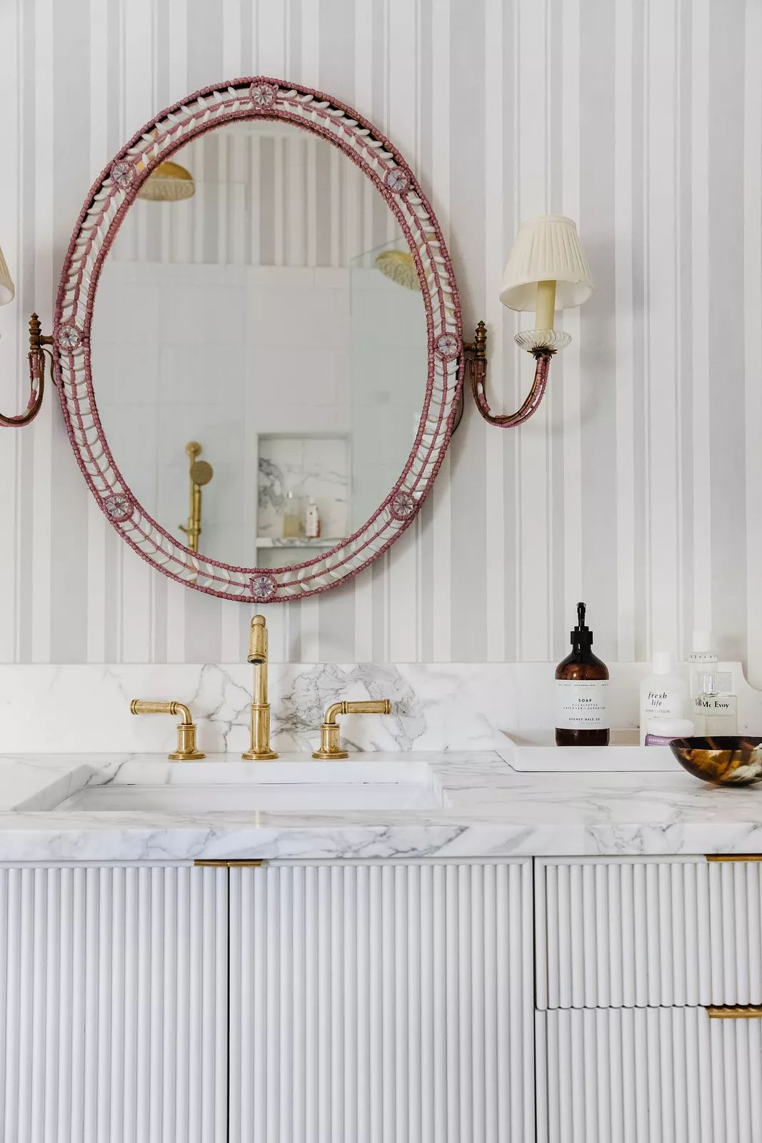 Bathroom with marble countertop, round mirror, wall sconces, ribbed cabinetry, and striped wallpaper.